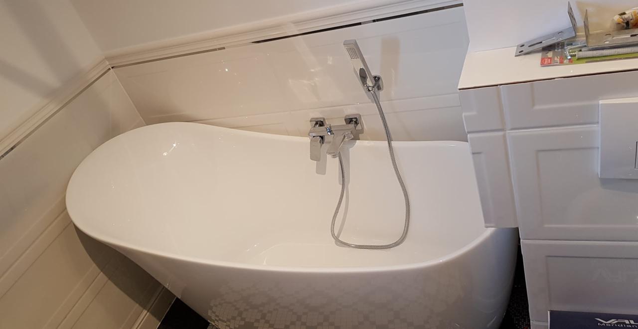 Wolnostojąca wanna z baterią i słuchawką prysznicową na tle białych płytek ściennych i zabudowy z widocznymi akcesoriami montażowymi.