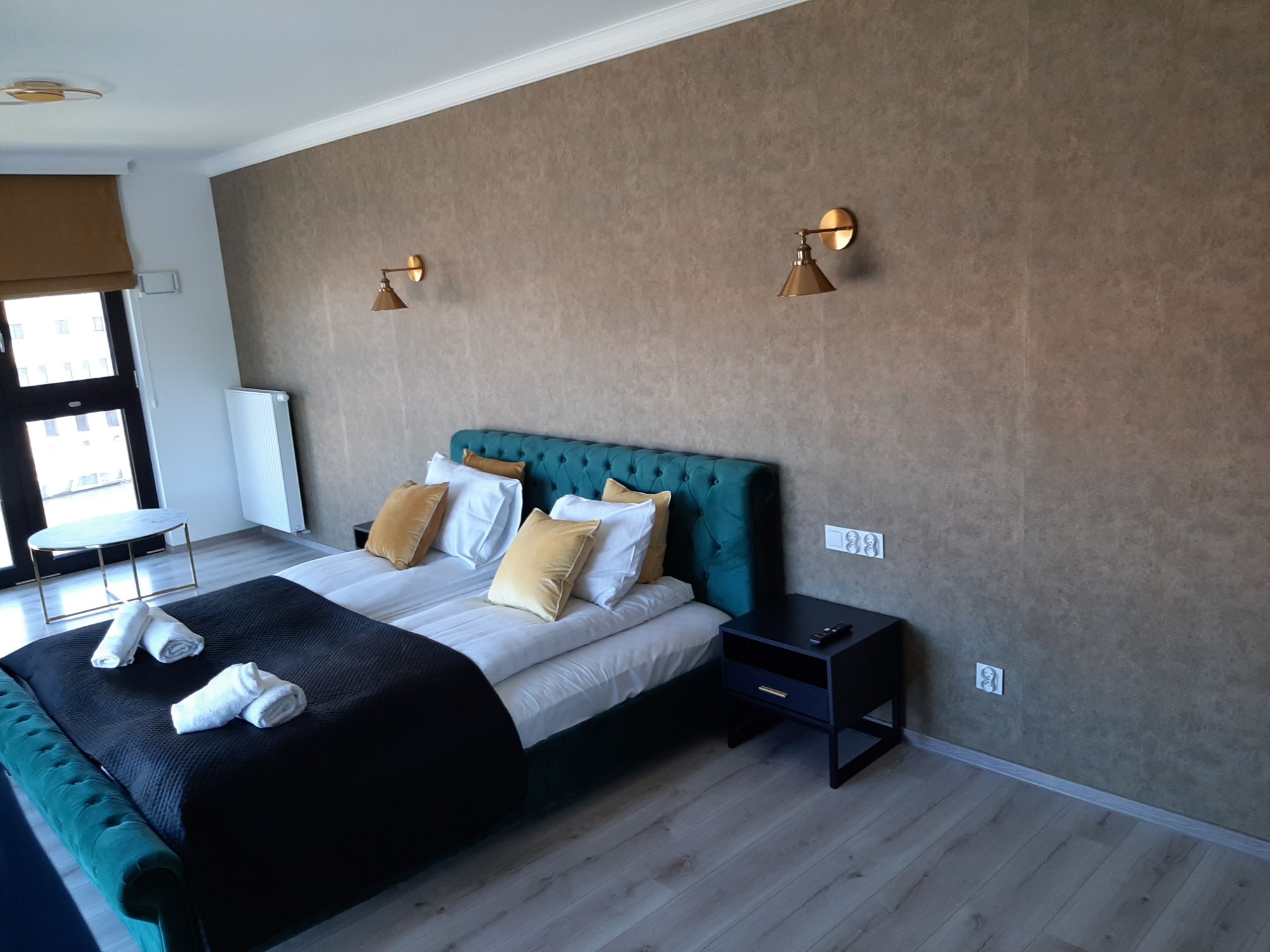 Nowoczesna sypialnia w Hopowie po wykończeniu, z łóżkiem z turkusowym zagłówkiem, złotymi lampami ściennymi i drewnianą podłogą.