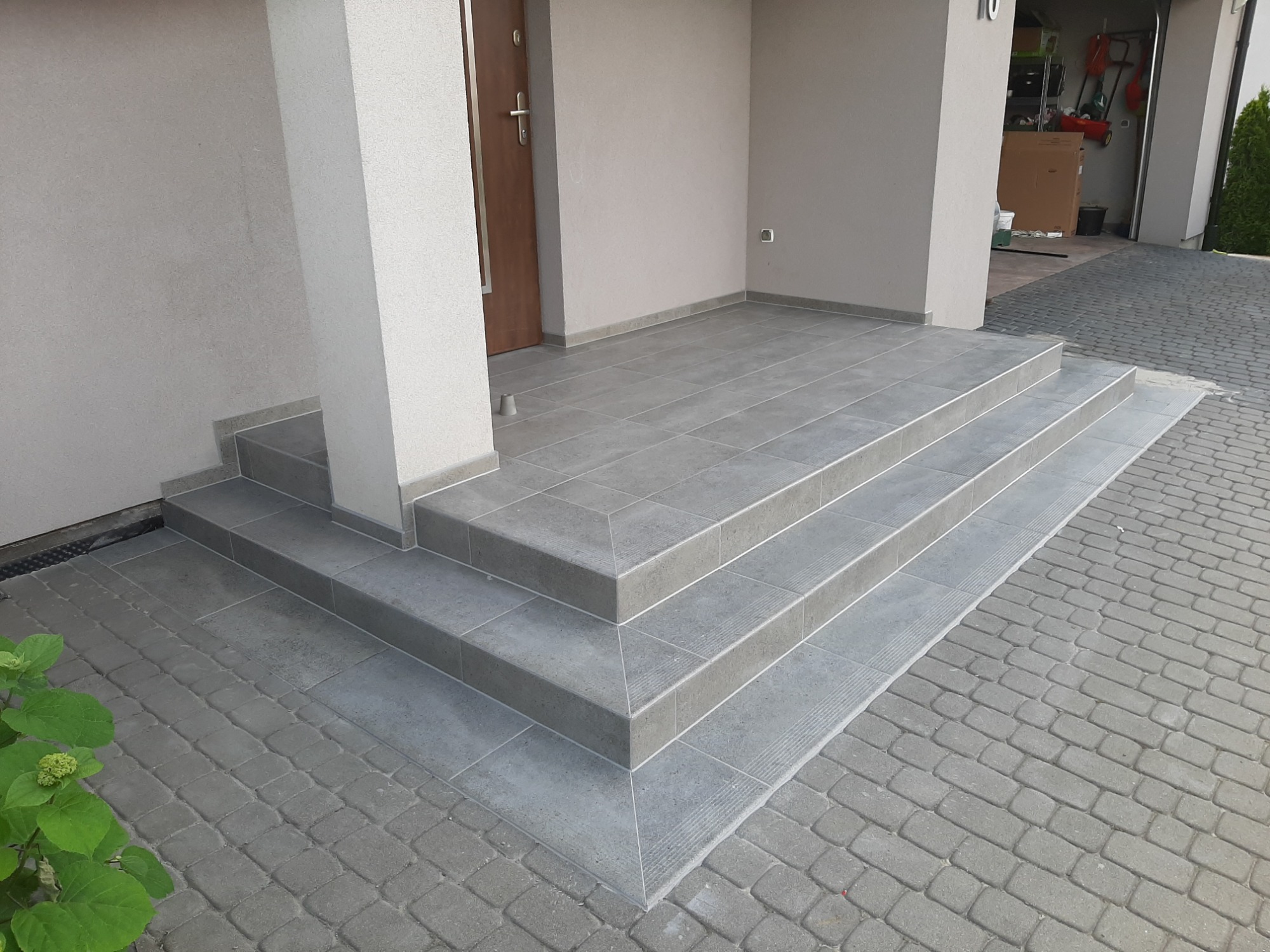 Zewnętrzne schody wejściowe do domu wyłożone szarą, kwadratową glazurą, z subtelnymi żłobieniami na krawędziach stopni, dopasowane kolorystycznie do kostki brukowej na podjeździe.