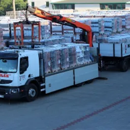 Rozładunek cegieł Roben z ciężarówki z przyczepą na placu składu budowlanego. Żuraw przenosi palety. Widoczny napis 'Materiały budowlane Wesołek'.