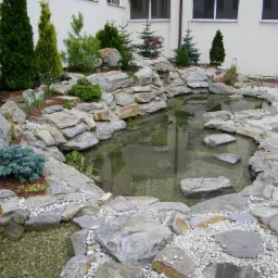 Oczko wodne z kamieni, otoczone roślinnością, z widocznym odbiciem budynku w wodzie.