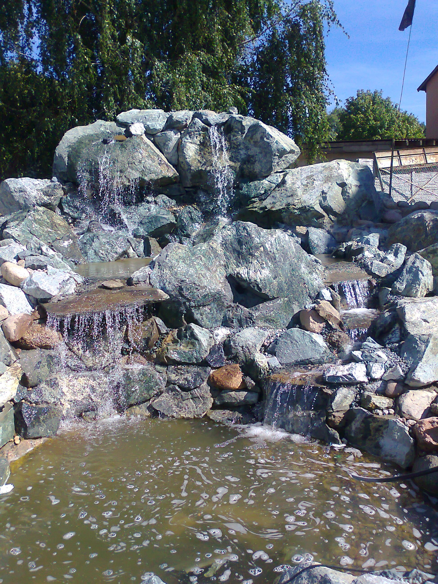 Kaskada wodna zbudowana z szarych i zielonych kamieni różnej wielkości, tworząca małe wodospady wpadające do stawu z lekko mętną wodą, na tle drzew i fragmentu budynku.