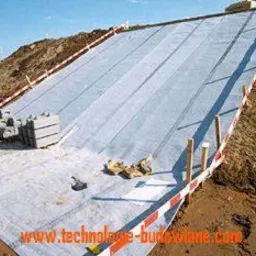 Rozłożona geowłóknina na zboczu, przygotowana pod dalsze prace budowlane, widoczne paliki z biało-czerwoną taśmą, robotnicy w tle i ułożone bloczki betonowe. W dolnej części zdjęcia widoczny adres...