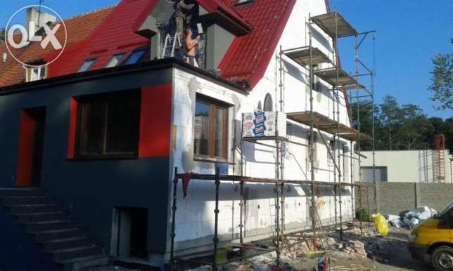 Ocieplanie domu z czerwonym dachem, widoczne rusztowania i pracownicy na dachu. Część elewacji pomalowana na szaro z czerwonymi akcentami.