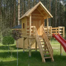 Drewniany domek zabaw dla dzieci z czerwoną zjeżdżalnią, schodami i małym balkonem, umieszczony na trawiastym terenie z widokiem na las.