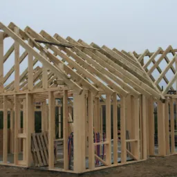 Konstrukcja szkieletowa drewnianego budynku w trakcie budowy, widoczne elementy dachu i ścian, na zewnątrz paleta i rury.