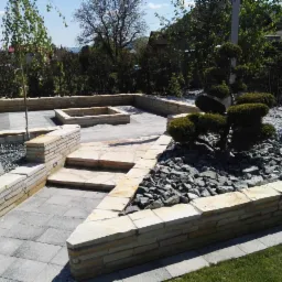 Ogr&oacute;d z kamienną architekturą: ścieżki z płyt, schody, murek z piaskowca, rabata z ozdobnymi kamieniami i formowana roślina, w tle drzewa i zabudowa.