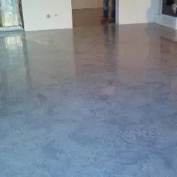 Gładka, szara posadzka z efektem polerowanego betonu w jasnym wnętrzu, widoczne odbicia światła i fragmenty instalacji grzewczej przy ścianie.