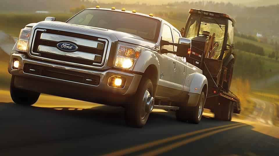 Laweta z traktorem w Szczecinie. Biały Ford Super Duty transportuje sprzęt budowlany na niskopodwoziowej przyczepie. Ujęcie z dołu, w tle pagórkowaty krajobraz.