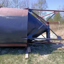 Metalowy silos z widocznymi śladami rdzy i świeżo nałożoną, ciemną farbą, oparty na drewnianym klocku na tle trawy i drzew.