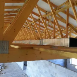 Wnętrze budynku w trakcie budowy z widoczną drewnianą konstrukcją dachu i systemem wentylacyjnym z metalowych kanałów.