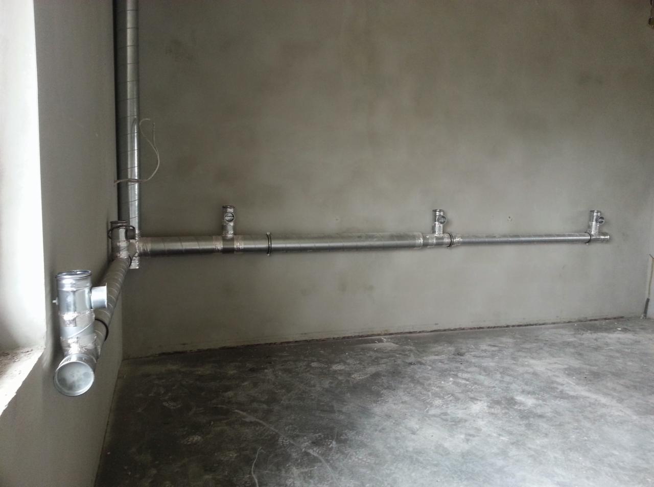 Szare pomieszczenie z zamontowaną, poziomą, metalową rurą z zaworami, połączoną z pionową rurą przy ścianie. Widoczne łączenia i fragment podłogi.
