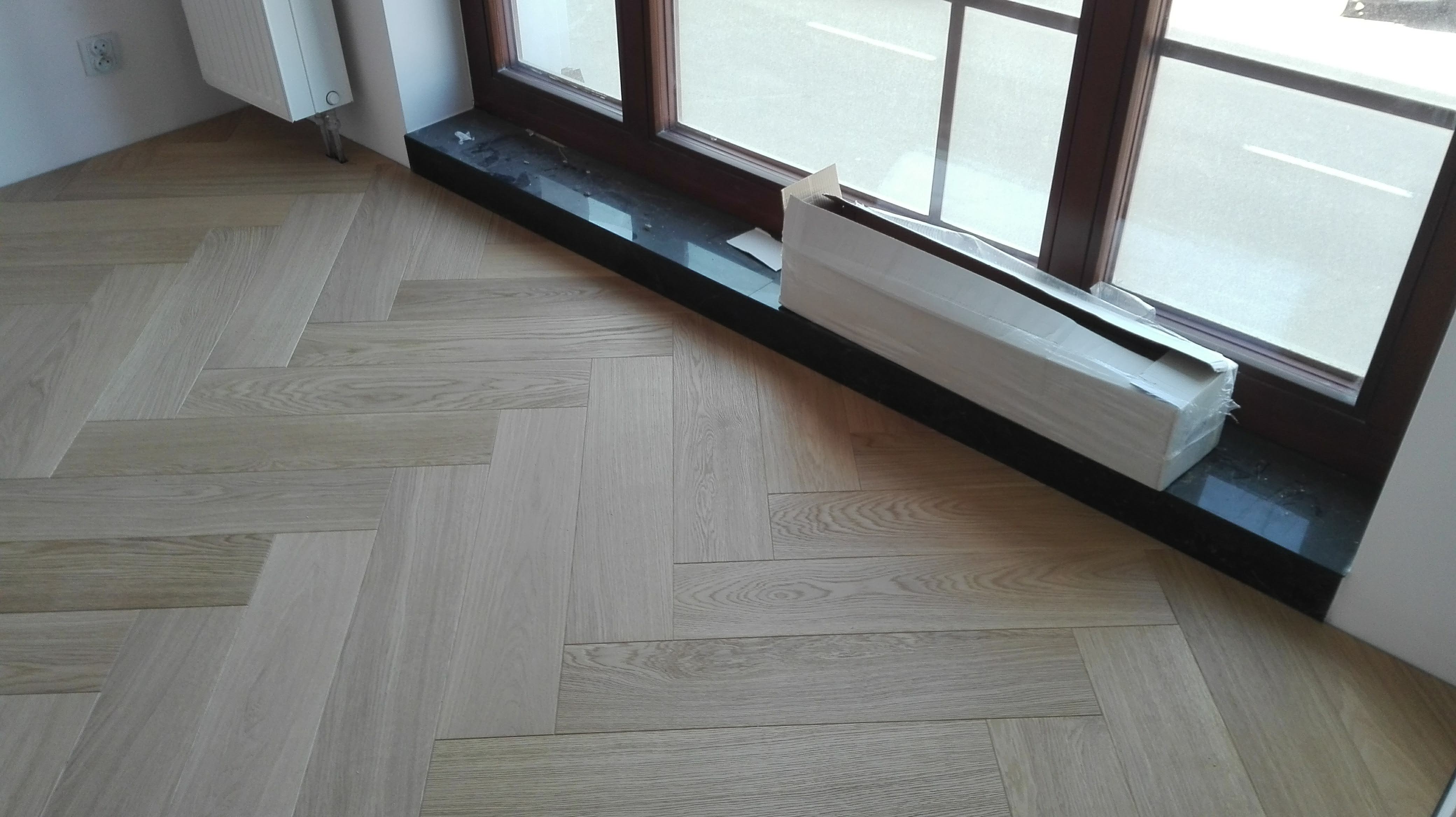 Ułożona jasna podłoga z desek w jodełkę francuską, z paczką paneli czekającą na montaż przy oknie.