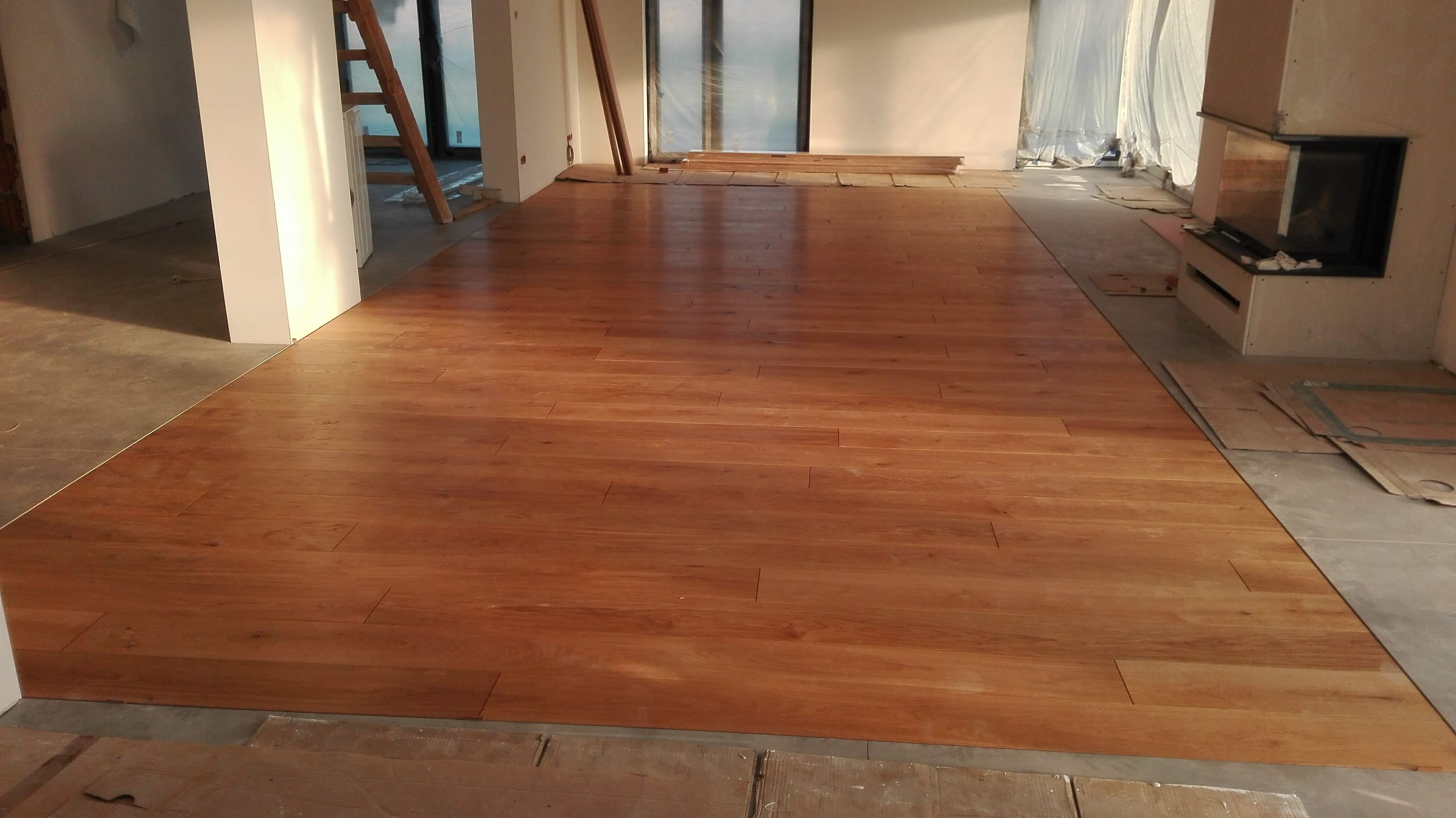 Ułożona, lakierowana podłoga z desek parkietowych w odcieniach brązu, widoczna w surowym wnętrzu z kominkiem i schodami, zabezpieczona kartonem przy ścianach.
