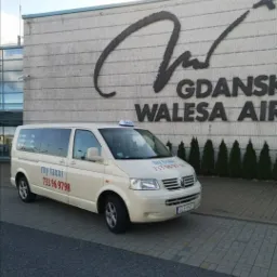 Jasny van taxi z logo 'my taxxi' i numerem telefonu 733 96 9798 na tle budynku lotniska Gdańsk Wałęsa Airport.