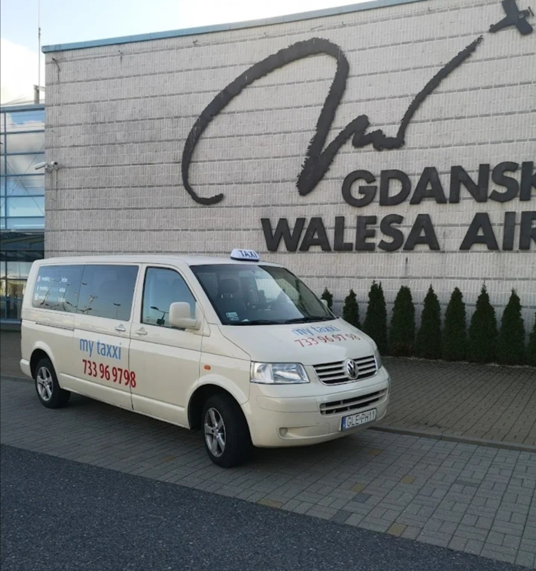 Jasny van taxi z logo 'my taxxi' i numerem telefonu 733 96 9798 na tle budynku lotniska Gdańsk Wałęsa Airport.