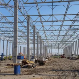 Wnętrze powstającej hali z widocznymi betonowymi słupami, stalową konstrukcją dachu i sprzętem budowlanym na placu budowy.