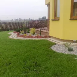 Aranżacja ogrodu z zielonym trawnikiem, rabatami z kostki i kamienia, obok żółtego domu z drewnianymi oknami, ogrodzenie w tle.