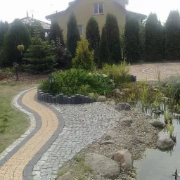 Kręta ścieżka z kostki brukowej w ogrodzie z oczkiem wodnym, obrzeża z palisady, w tle dom i wysokie tuje.