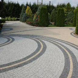 Granitowa nawierzchnia z dekoracyjnymi, łukowatymi pasami w odcieniach szarości i beżu, otoczona zielenią drzew i krzewów. Widok na podjazd.
