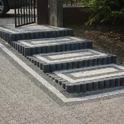 Schody z kostki brukowej w odcieniach szarości, prowadzące do furtki. Elementy dekoracyjne z jasnej kostki. Widoczne tło z zielenią i fragment auta.