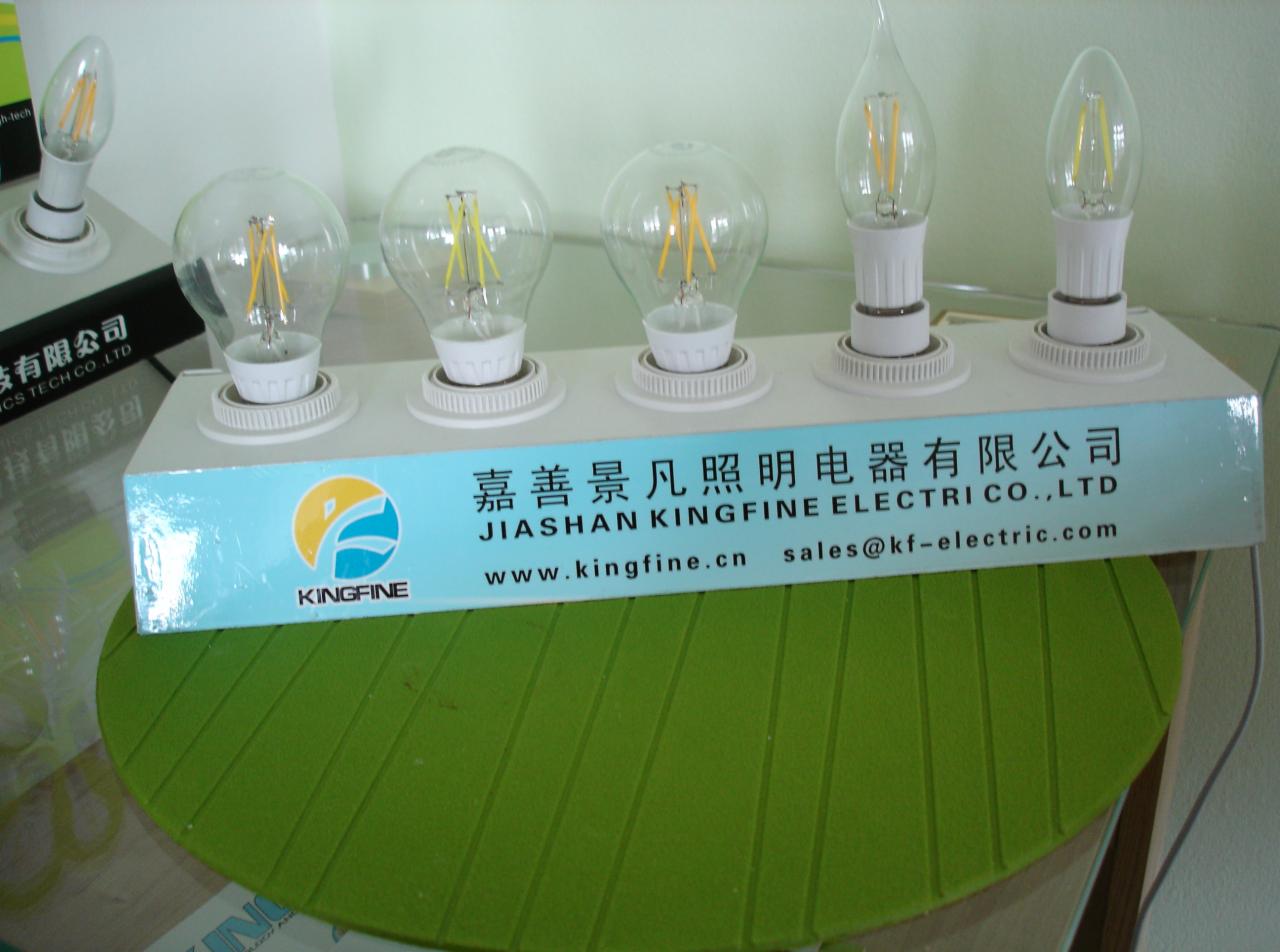 Różne typy żarówek LED na białej listwie z logo i adresem firmy Kingfine, na tle zielonego podkładu. Prezentacja asortymentu oświetlenia.