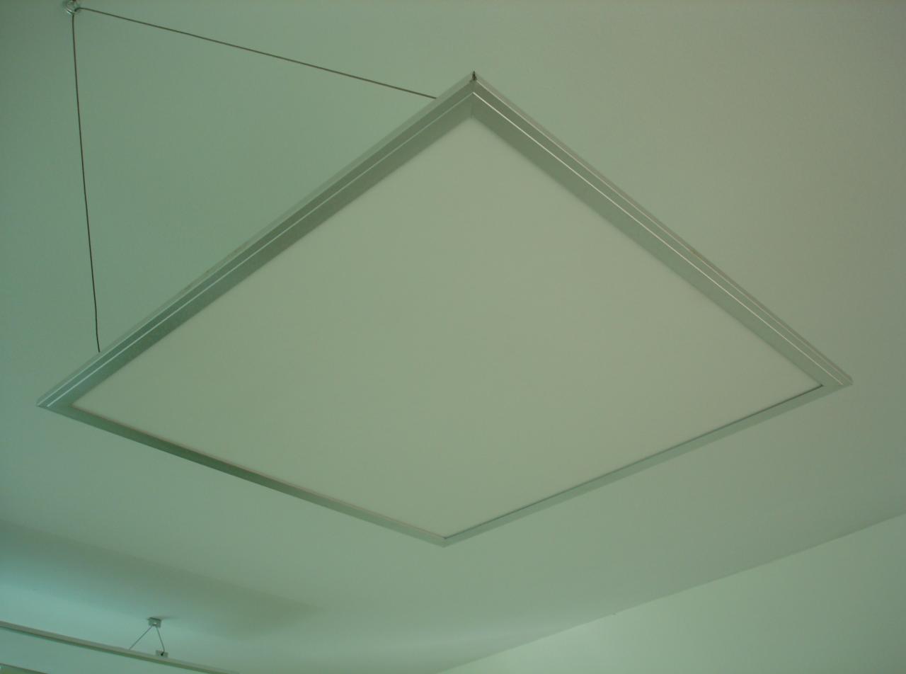 Kwadratowa lampa LED z aluminiową ramą, zawieszona na linkach, oświetla sufit. Minimalistyczny design.