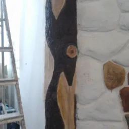 Fragment elewacji z dekoracyjnym tynkiem imitującym kamień i elementami w kolorze ciemnego brązu, przypominającymi korę drzewa, widoczna drabina malarska.