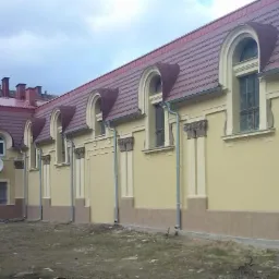 Odnowiona elewacja budynku z oknami w kształcie łuków, z nową czerwoną dachówką i elementami dekoracyjnymi w stylu klasycystycznym, widok z zewnątrz na tle pochmurnego nieba.