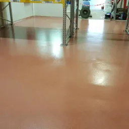 Gładka, brązowo-pomarańczowa podłoga w hali magazynowej z metalowymi regałami, widoczne ślady po pracy w tle.