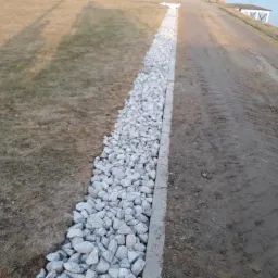 Wykonany rów wypełniony białym kamieniem, oddzielający trawnik od drogi gruntowej z widocznymi śladami opon, w tle budynek.