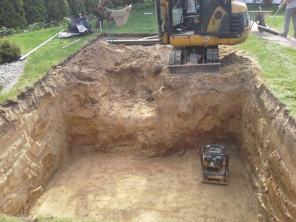 Wykop pod fundamenty domu jednorodzinnego z użyciem minikoparki i ubijarki wibracyjnej, widok z góry, trawa i zieleń w tle.