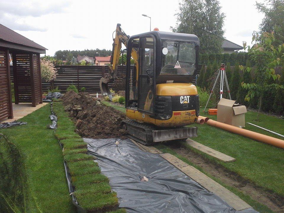 Żółta minikoparka CAT podczas prac ziemnych w ogrodzie, widoczne wykopane koryto, rury i trawa w rolkach. Niwelator w tle. Prace przygotowawcze pod instalację.