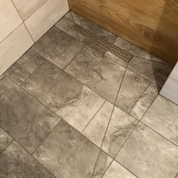 Szare płytki podłogowe w łazience z odpływem liniowym z motywem greckim, widoczne fragmenty drewnopodobnej okładziny ściennej i jasnych płytek.