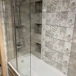 Nowoczesna łazienka z wanną i szklanym parawanem, ściany wyłożone szarymi i wzorzystymi płytkami, chromowana bateria prysznicowa z deszczownicą.