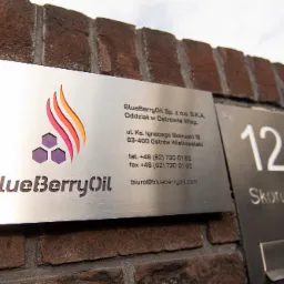 Tabliczka informacyjna firmy BlueberryOil z logo i adresem, umieszczona na ceglanej ścianie obok domofonu z numerem 12.