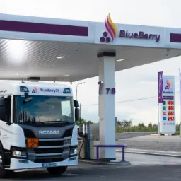 Cysterna z logo BlueBerryOil na stacji paliw z wyświetlaczem cen i flagami reklamowymi, w tle zieleń.