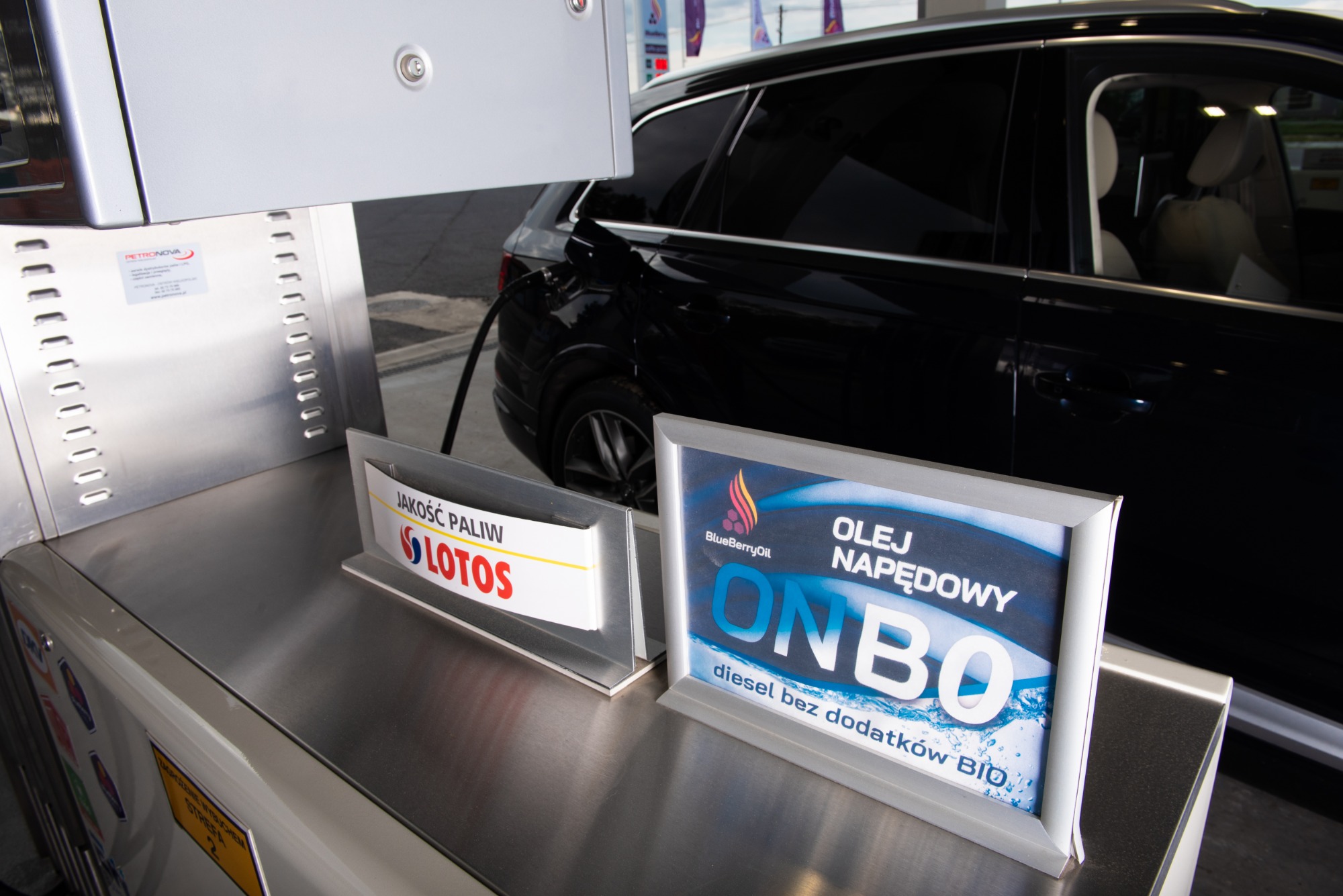Tankowanie czarnego samochodu na stacji paliw, widoczne oznaczenia 'Jakość Paliw Lotos' oraz informacja o 'Oleju Napędowym ONBO diesel bez dodatków BIO'.