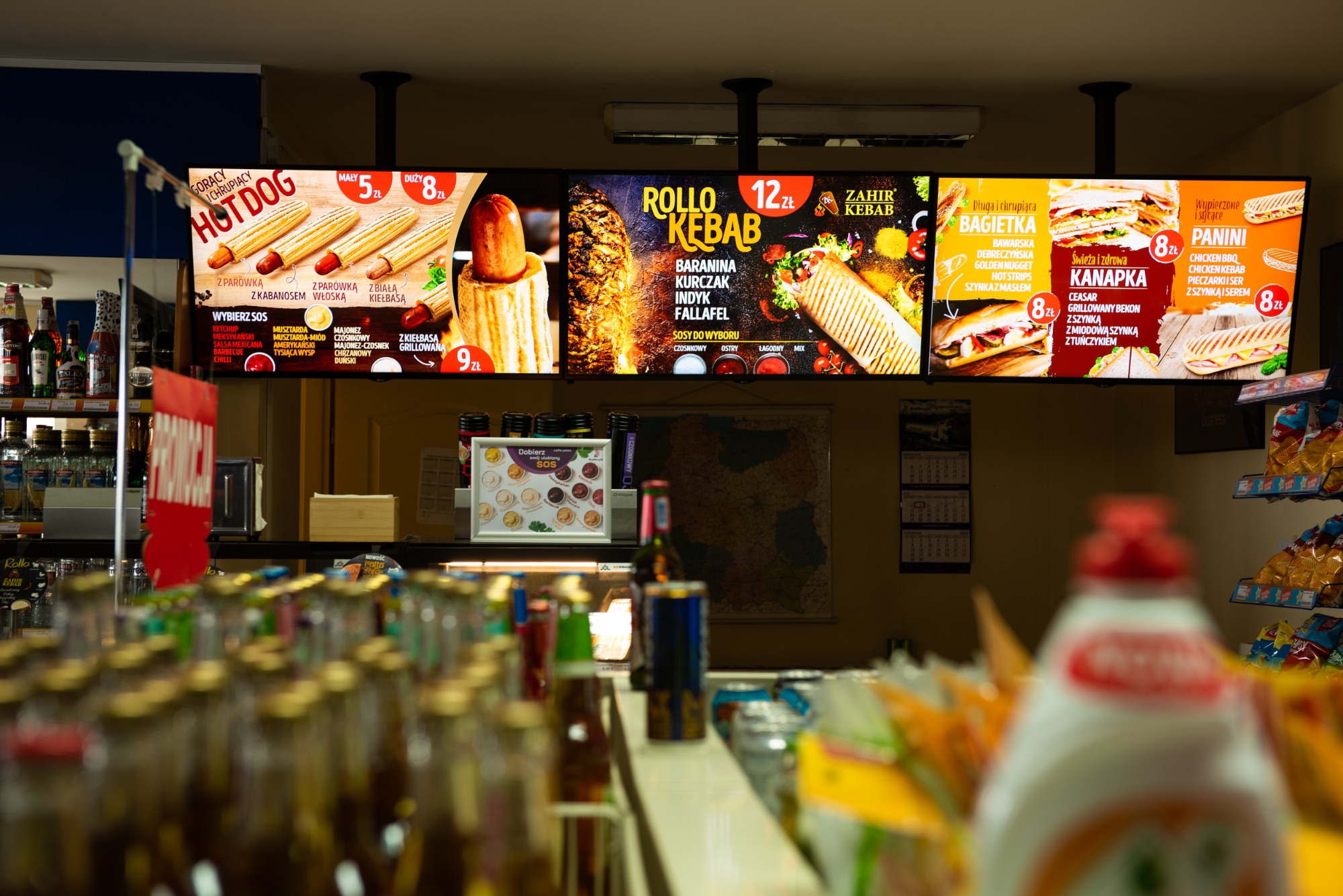 Podświetlane menu fast food z hot dogami, kebabami, bagietkami i kanapkami nad ladą chłodniczą z napojami i przekąskami w sklepie.