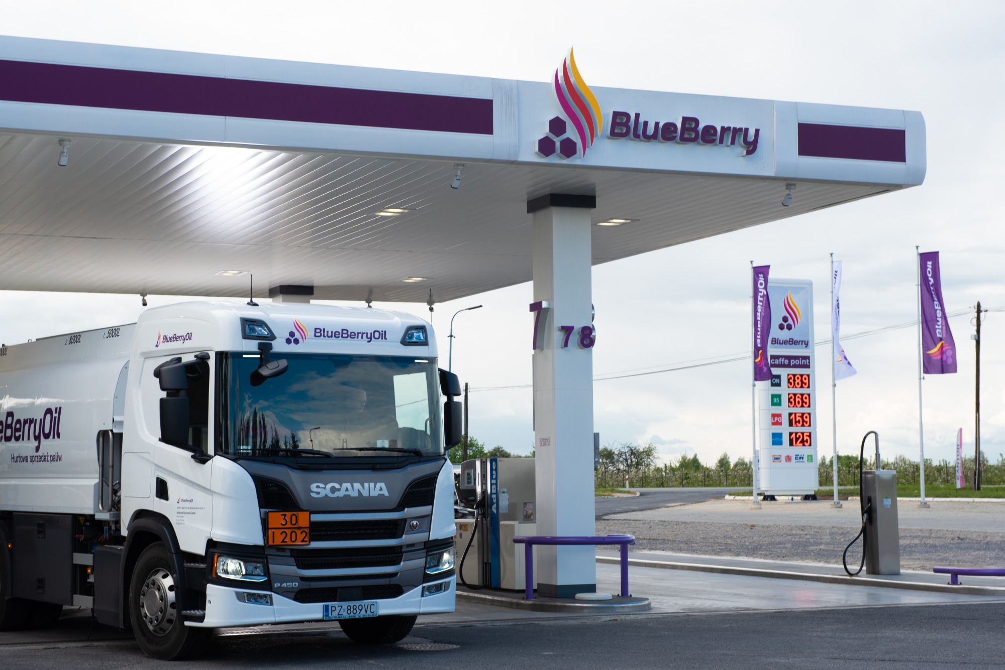 Cysterna z logo BlueBerryOil na stacji paliw z wyświetlaczem cen i flagami reklamowymi, w tle zieleń.