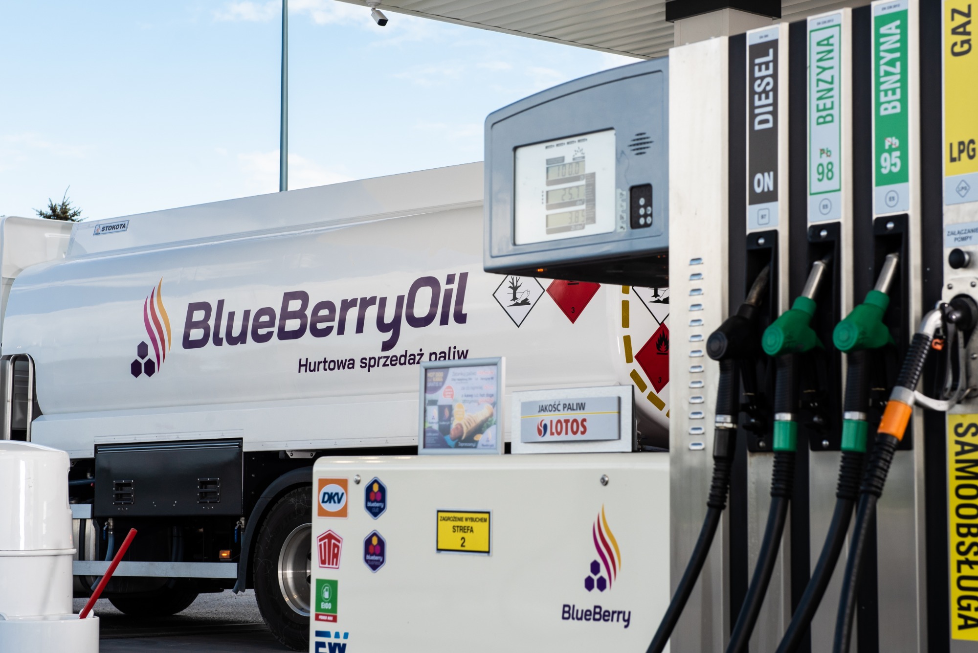Cysterna z logo BlueBerryOil na stacji paliw, dystrybutory z pistoletami do diesla, benzyny 98 i 95 oraz LPG, tabliczka informacyjna o jakości paliw Lotos.