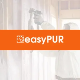 Pracownik w kombinezonie ochronnym aplikuje pianę izolacyjną na drewnianą konstrukcję ściany wewnętrznej za pomocą pistoletu natryskowego; widoczny pomarańczowy baner z logo firmy.