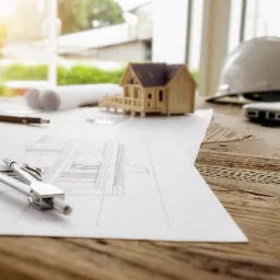 Projekt domu na desce kreślarskiej z cyrklem, ołówkiem, modelem domu, kaskiem budowlanym i laptopem w tle, oświetlone promieniami słonecznymi.