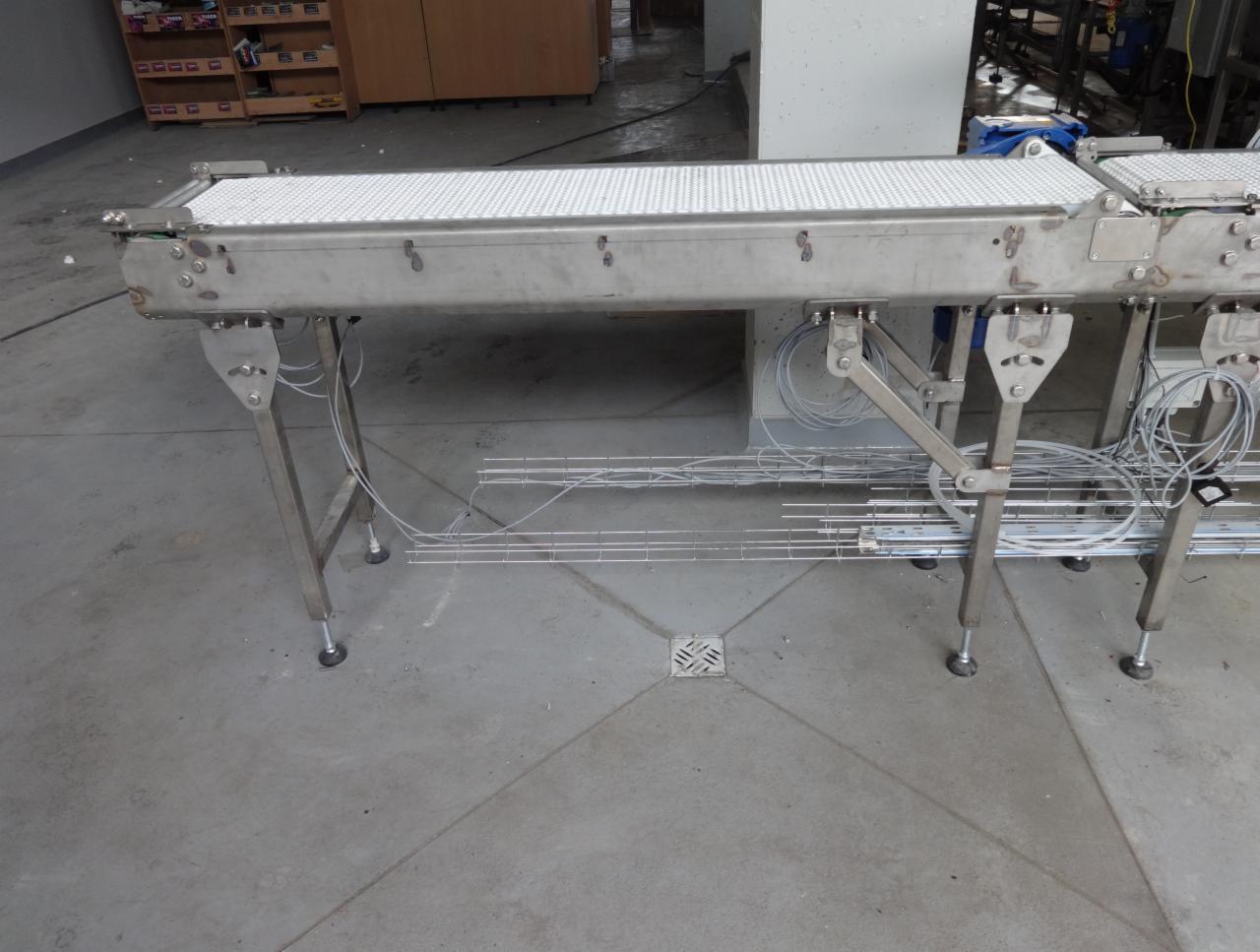 Metalowy przenośnik taśmowy z białym pasem, widoczne przewody i elementy montażowe, ustawiony na szarym betonowym podłożu.