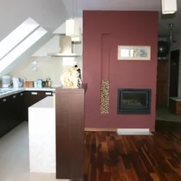 Nowoczesna kuchnia w poddaszu z ciemnymi szafkami, białym blatem i wyspą, widok na salon z ciemną ścianą z kominkiem i drewnem ułożonym w pionowej wnęce.