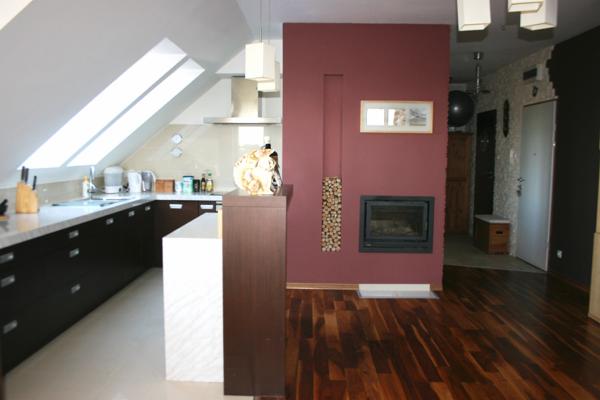 Nowoczesna kuchnia w poddaszu z ciemnymi szafkami, białym blatem i wyspą, widok na salon z ciemną ścianą z kominkiem i drewnem ułożonym w pionowej wnęce.