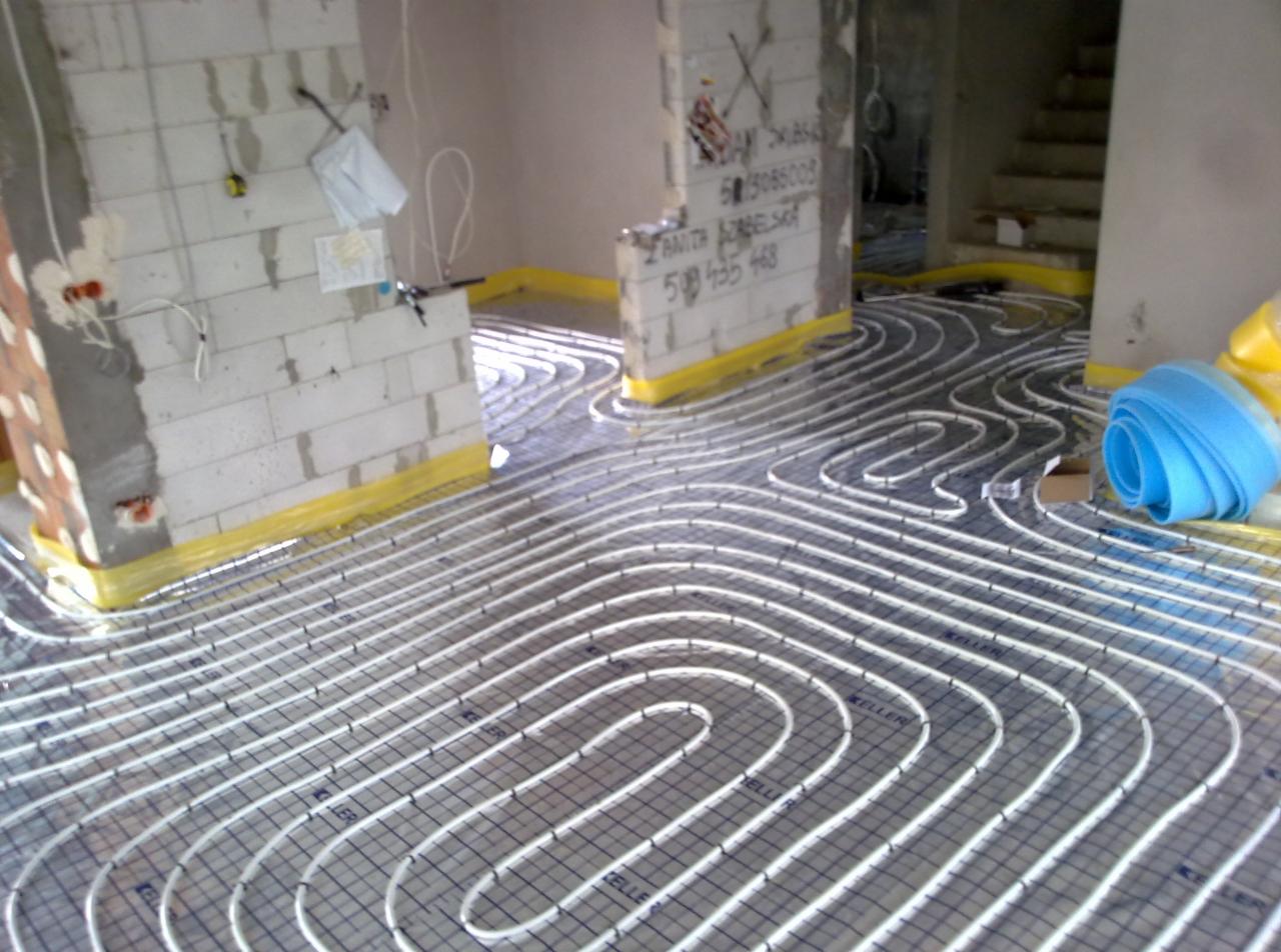 Białe rury ogrzewania podłogowego ułożone na folii izolacyjnej w surowym wnętrzu, zabezpieczone żółtą taśmą przy ścianach z betonu komórkowego.