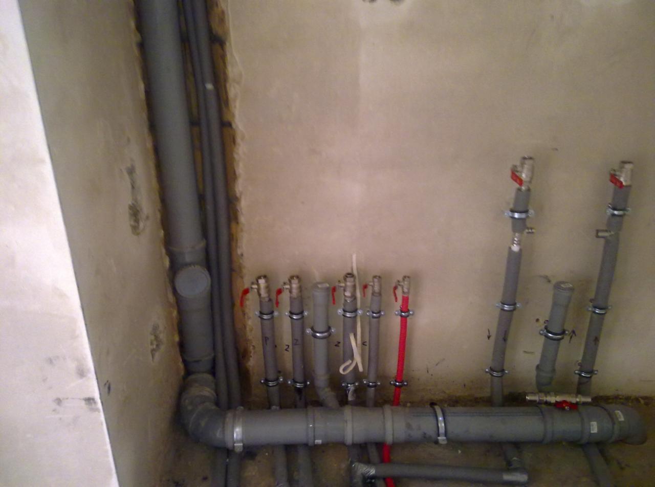 Szare i czerwone rury instalacji wodnej z zaworami, zamocowane do ściany klamrami. Widoczne złącza i fragmenty ścian w surowym stanie.