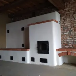 Biały piec kaflowy z ceglanymi akcentami i czarnymi drzwiczkami na tle ściany z cegły i drewnianego stropu.