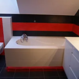 Nowoczesna łazienka na poddaszu z czarno-czerwonym akcentem na płytkach ściennych, białą wanną obudowaną płytkami, podświetleniem LED u dołu wanny i czarną podłogą z płytek.